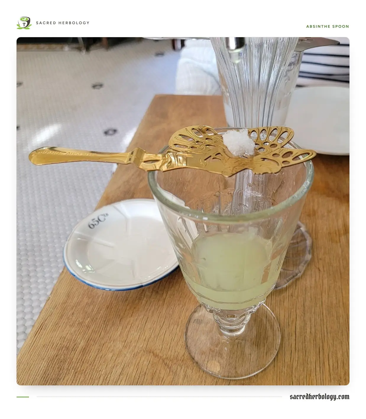 A pierced gilt absinthe spoon with a sugar cube resting across a glass of cloudy pale green absinthe, water dripping from a glass fountain behind
