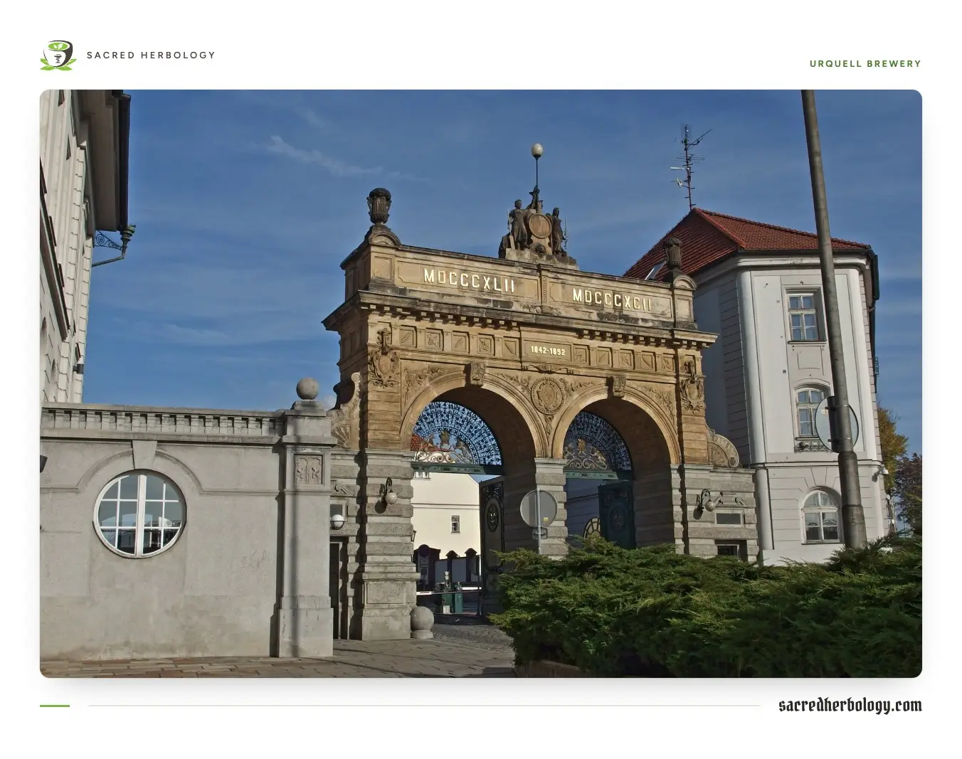 A sandstone double-arched gateway topped with statues and the dates 1842 and 1892, at the entrance to the Pilsner Urquell brewery under a blue sky