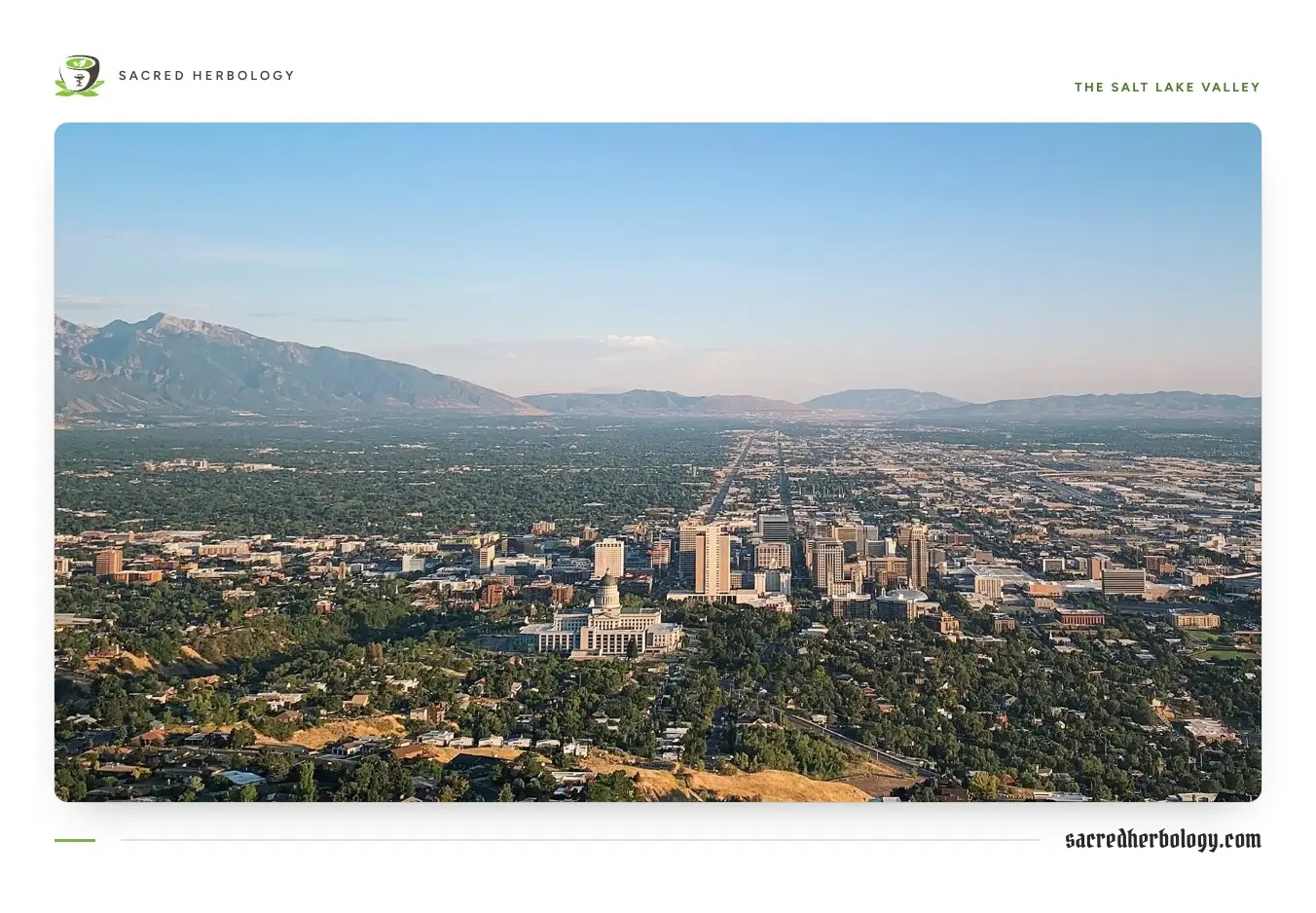 View over a wide green city in a valley, with a domed capitol building and downtown towers in the foreground and a long mountain range on the horizon