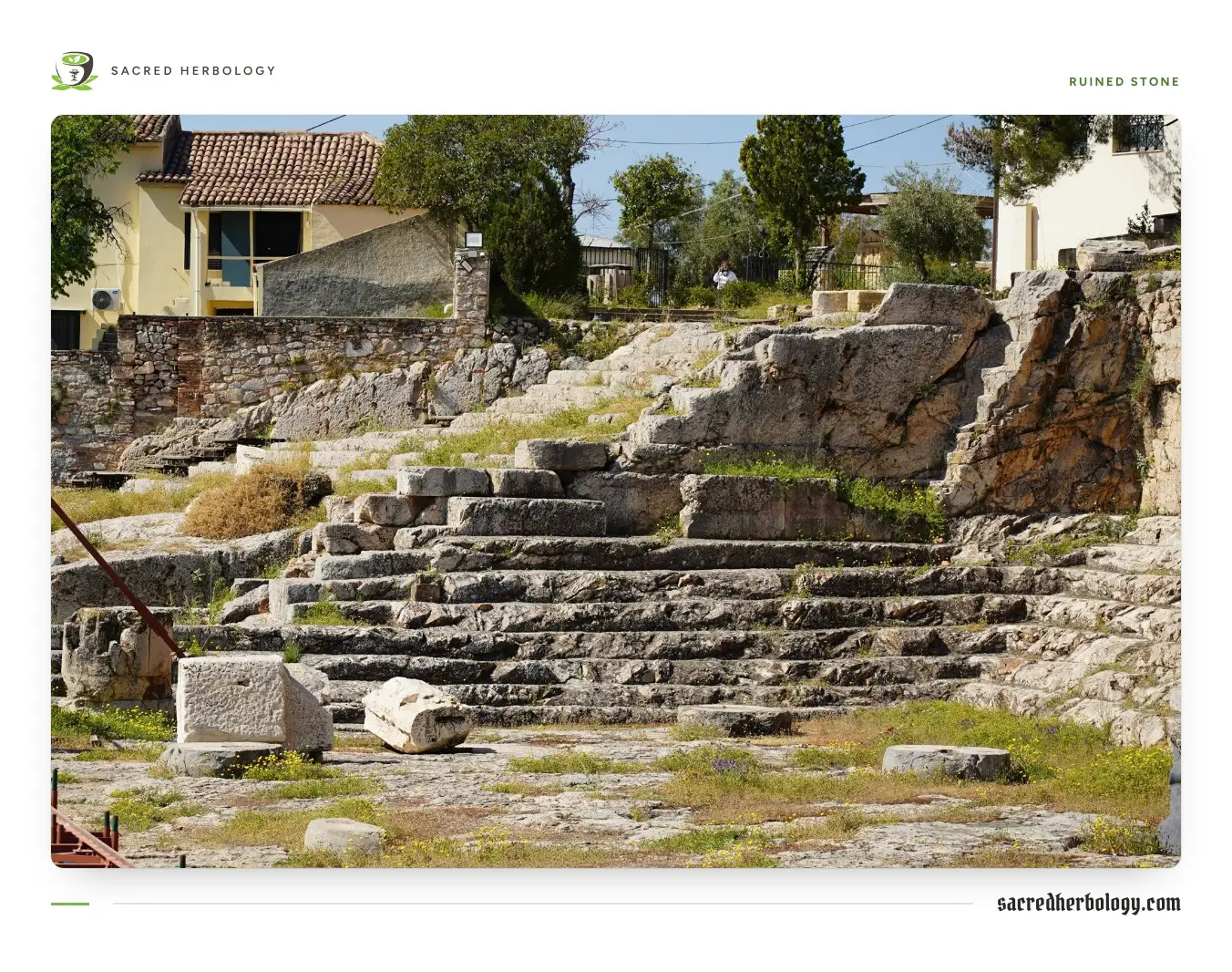 Ruined stone steps cut into a rocky slope at an archaeological site, with grass growing between scattered blocks and houses beyond